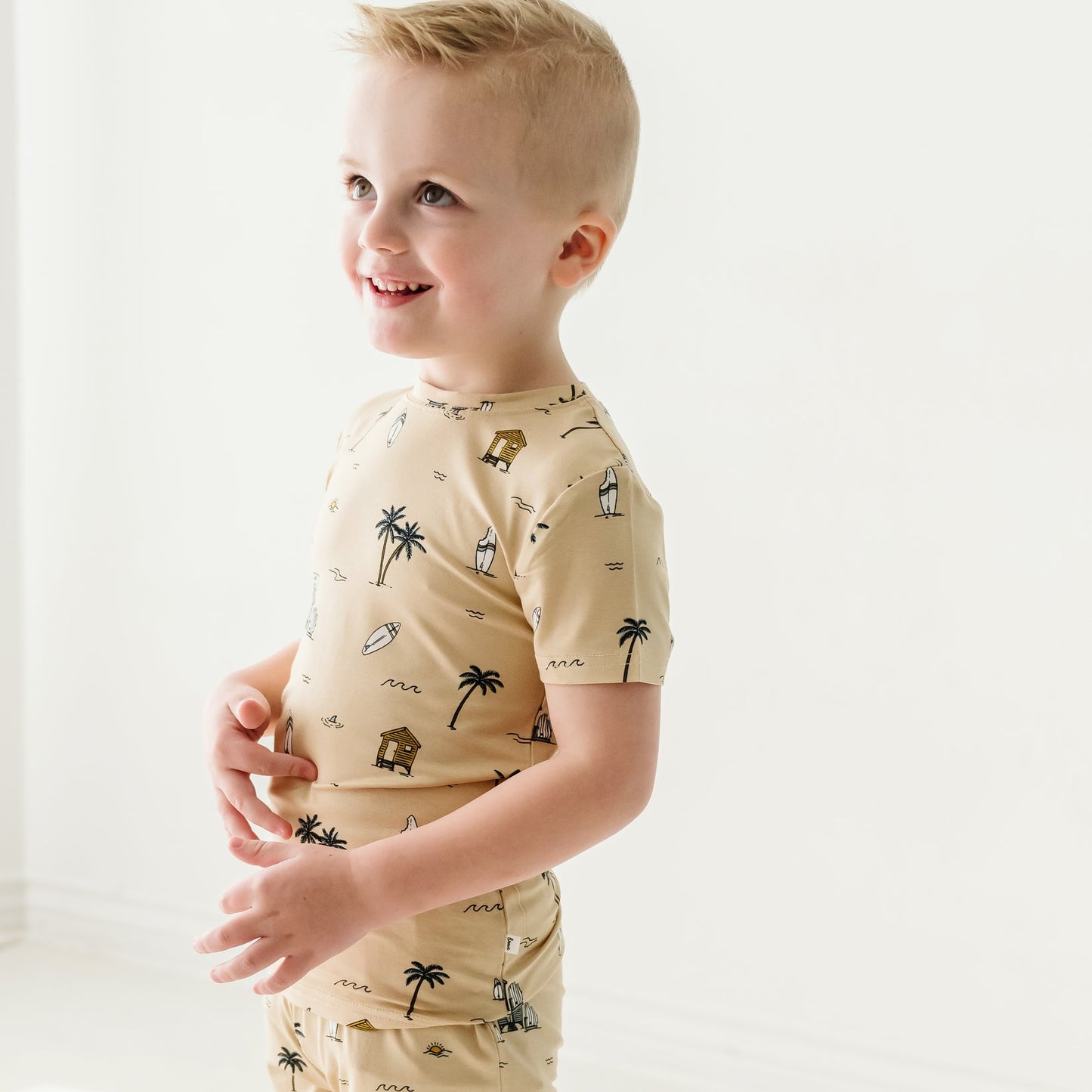 
                  
                    Side view of young boy in tan beach-print two-piece outfit featuring huts, waves, and palm trees – Surf Shack summer collection.
                  
                