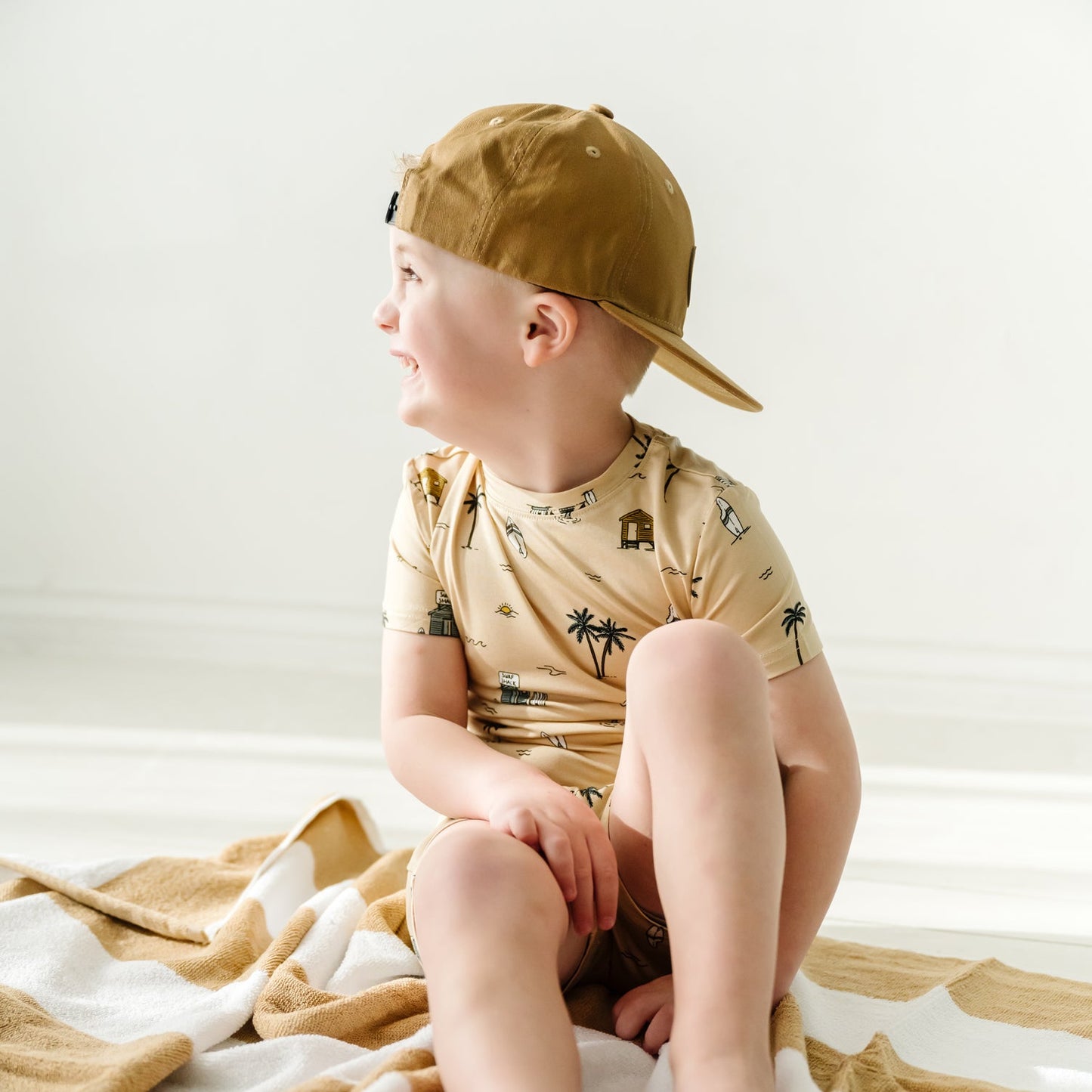 
                  
                    Little boy in Surf Shack shorts set sitting on striped towel, wearing tan backward cap and looking off to the side.
                  
                