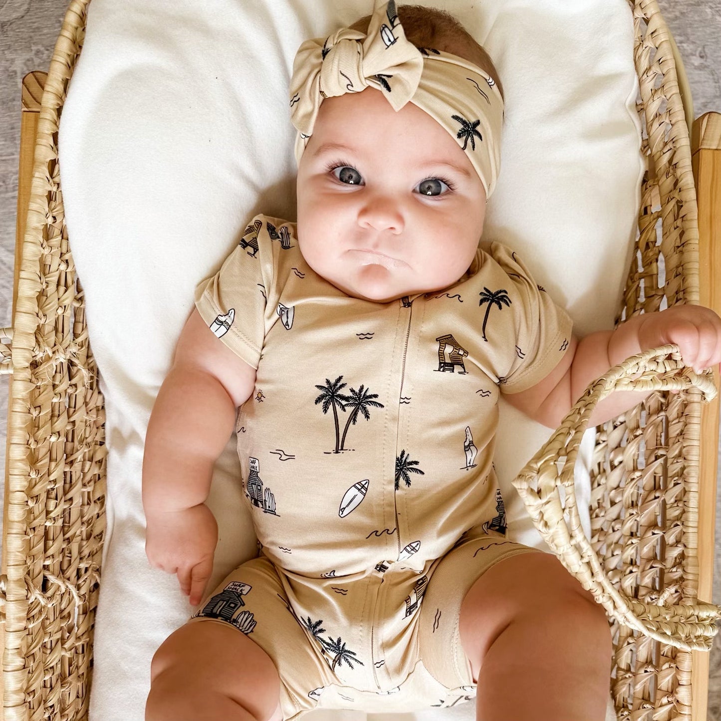 
                  
                    Baby girl wearing tan zip romper and matching bow headwrap with Surf Shack print, lying in wicker bassinet.
                  
                